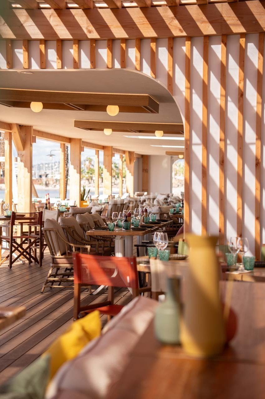 Terrasse d'un restaurant avec mobilier en bois, tables dressées et lumière naturelle filtrée par une pergola.
