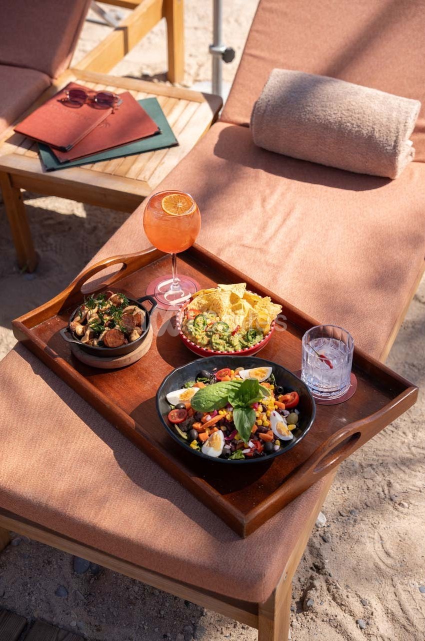 Plateau repas avec salade, guacamole, chips, légumes grillés et boisson, posé sur un transat en bord de plage.