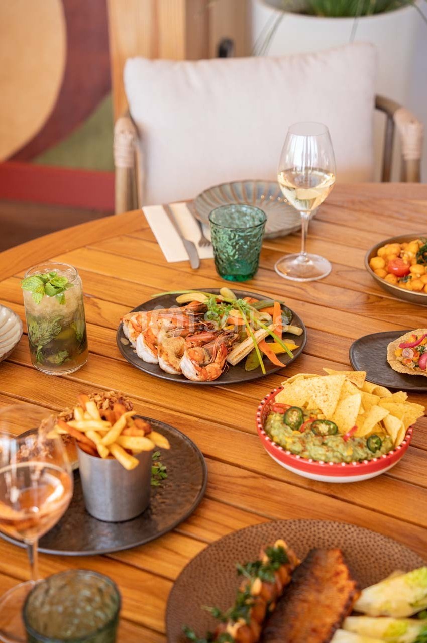 Table en bois avec des plats variés : fruits de mer, frites, nachos, guacamole, boissons et verres de vin.