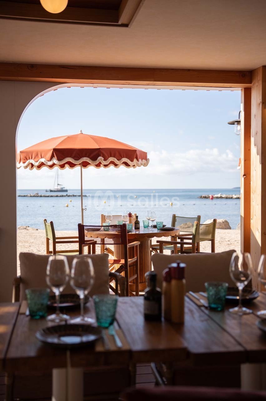 Vue sur une terrasse de restaurant en bord de mer avec des tables dressées et un parasol orange.