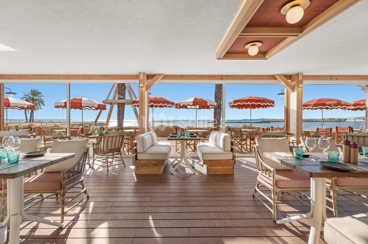 Terrasse d'un restaurant en bord de mer avec mobilier en bois, parasols rouges et vue sur la plage en arrière-plan.