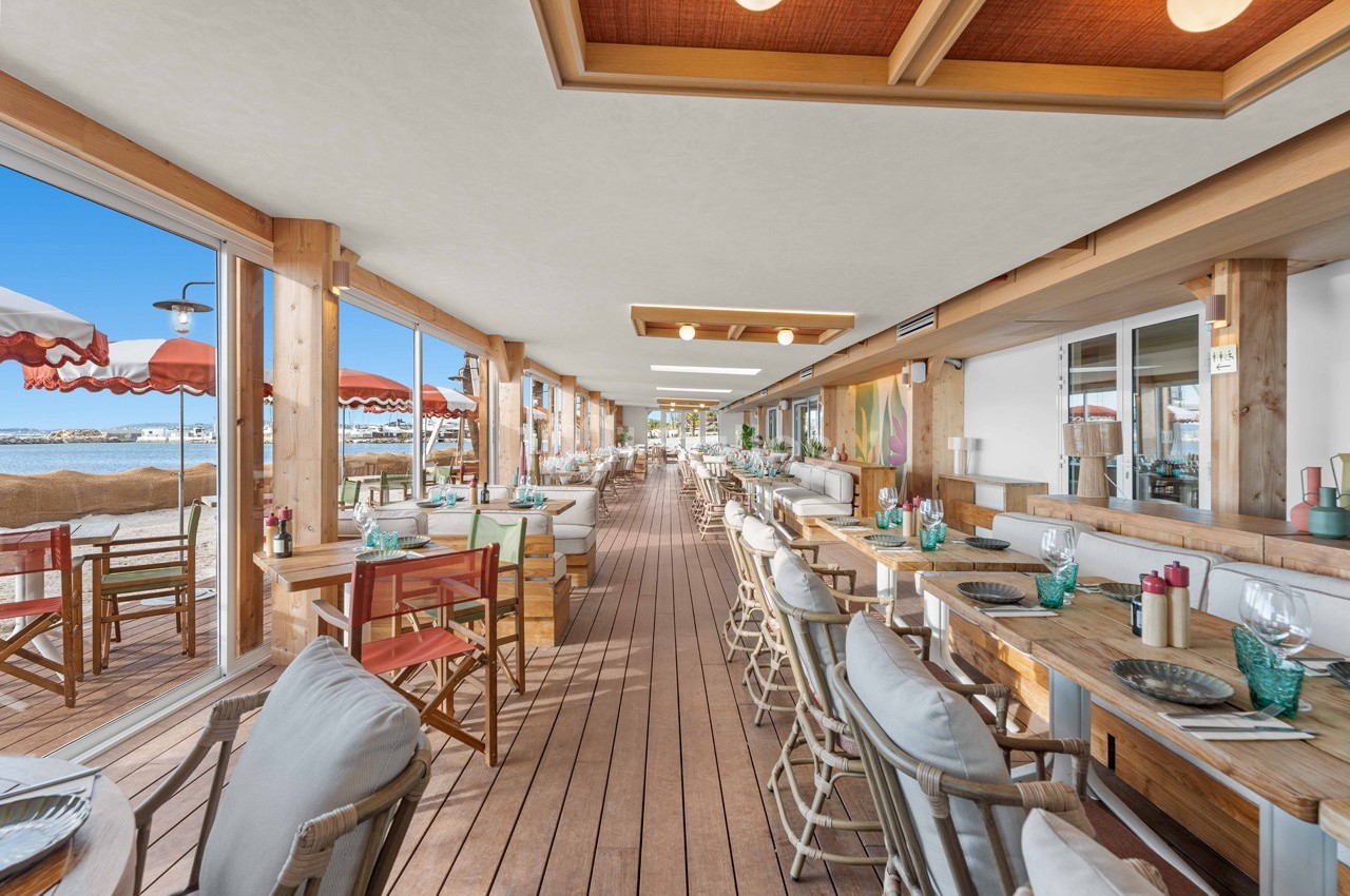 Salle de restaurant en bord de plage avec tables en bois, chaises confortables et vue sur la mer.