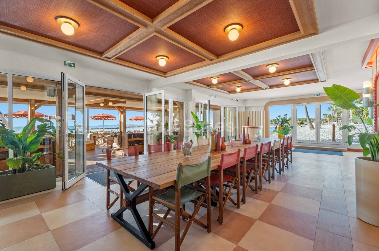 Salle lumineuse avec une grande table en bois, chaises colorées, plantes et vue sur une terrasse extérieure avec parasols…