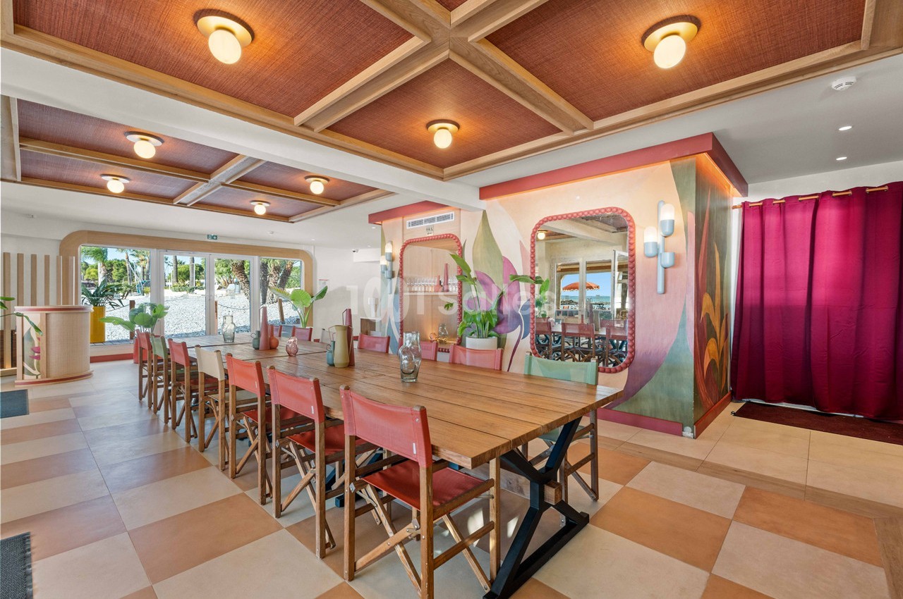Salle à manger lumineuse avec grande table en bois, chaises rouges et décorations colorées sur les murs.