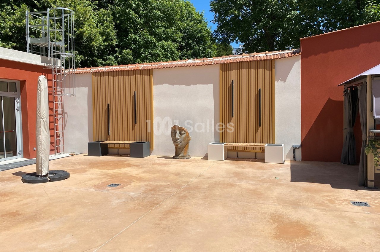 Cour extérieure avec mur blanc et orange, bancs en bois, sculpture de tête et échelle métallique à gauche.