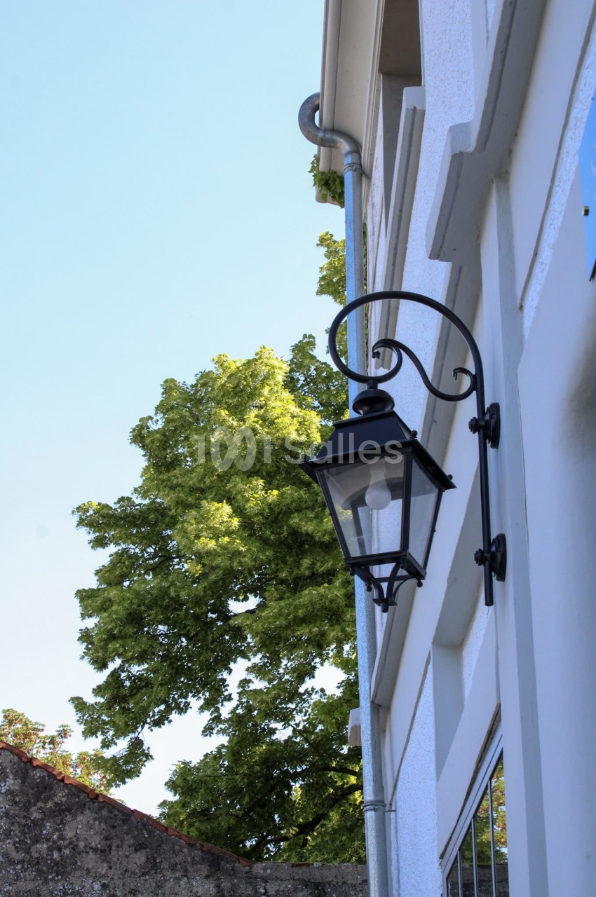 Lampe murale noire fixée sur une façade blanche, avec un arbre et un ciel bleu en arrière-plan.