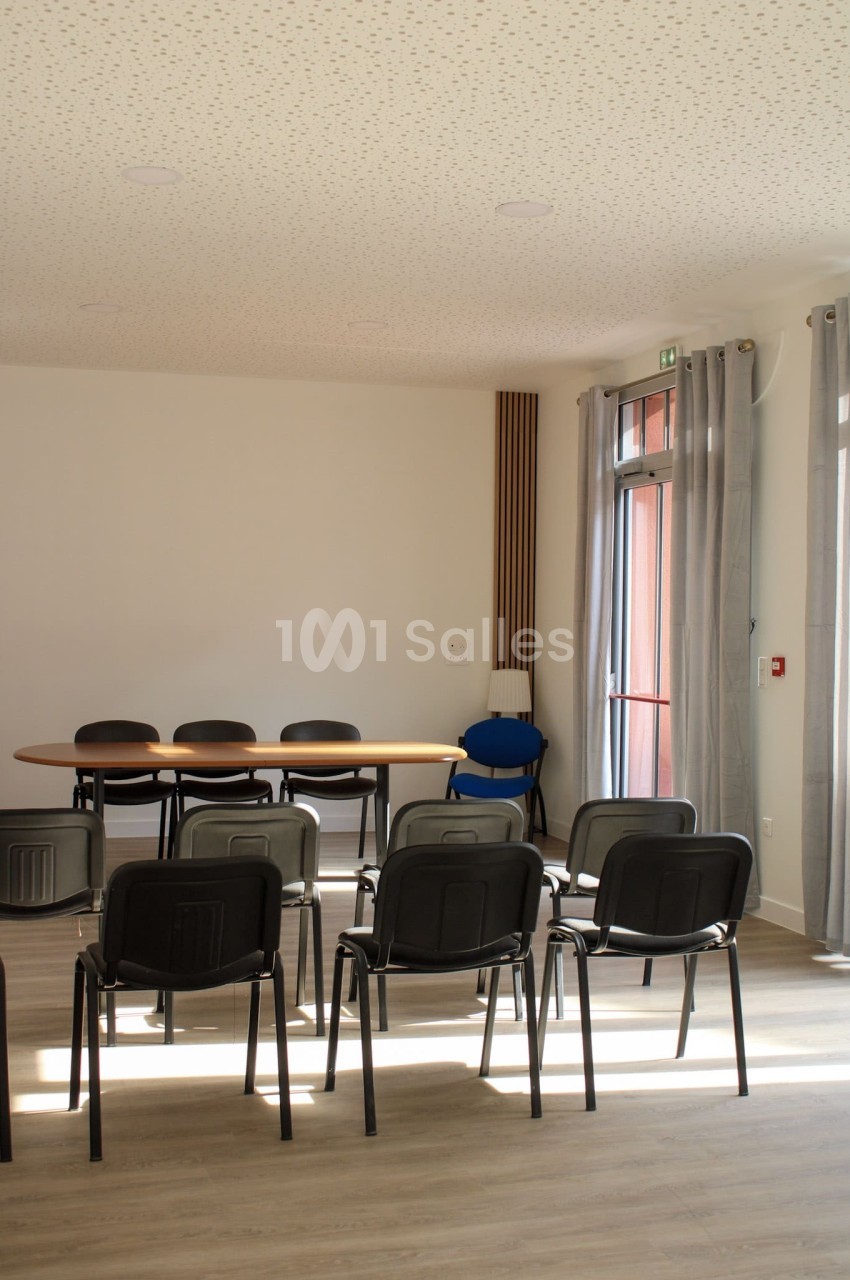 Salle de réunion lumineuse avec des chaises alignées face à une table ovale, près de grandes fenêtres avec rideaux.
