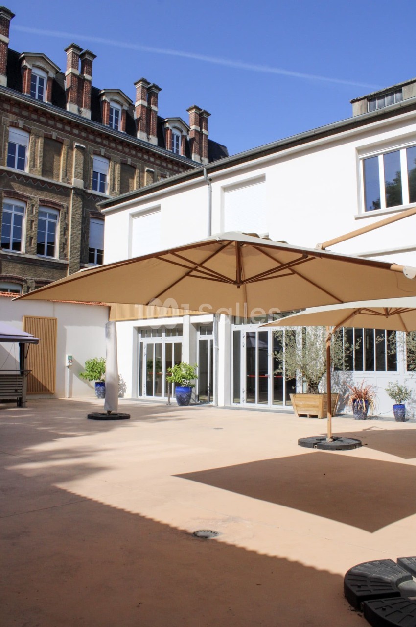 Cour extérieure ensoleillée avec parasols, sol beige, plantes en pots et bâtiments en arrière-plan.