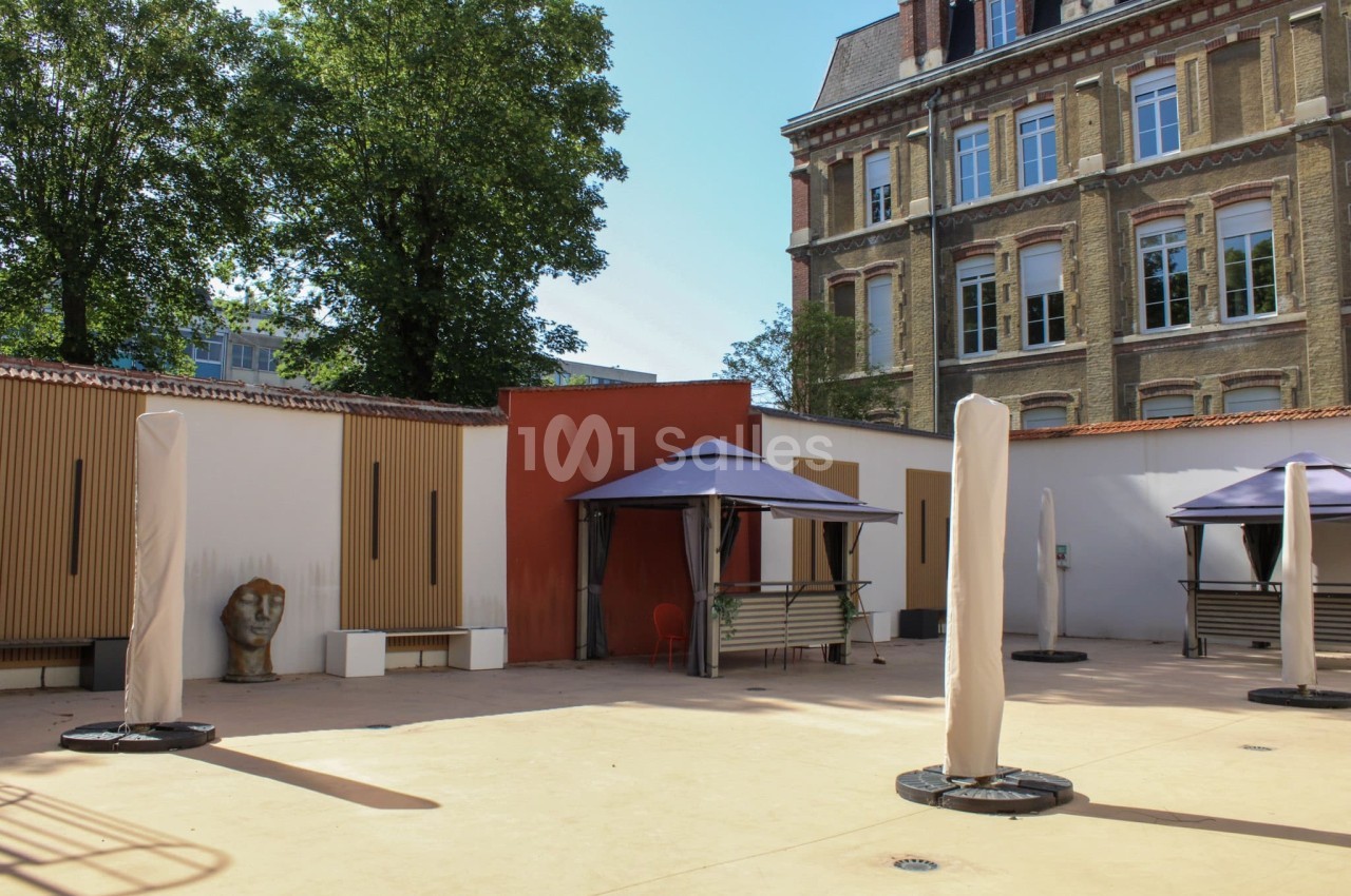 Cour extérieure avec sol en béton, parasols pliés, bancs, et bâtiments en briques avec fenêtres en arrière-plan.