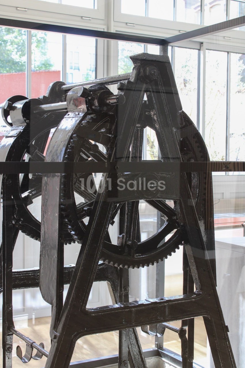 Machine industrielle ancienne en métal noir, exposée dans un espace lumineux avec de grandes fenêtres en arrière-plan.