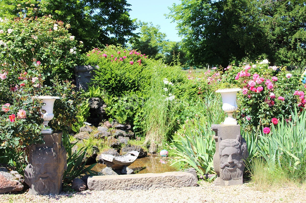 Jardin fleuri avec rosiers, statues sculptées et petit ruisseau entouré de verdure.