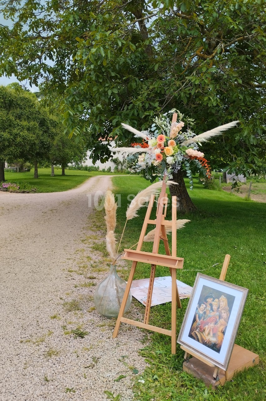 Chevalet en bois avec bouquet floral et tableau, disposé près d'un chemin gravillonné dans un jardin verdoyant.