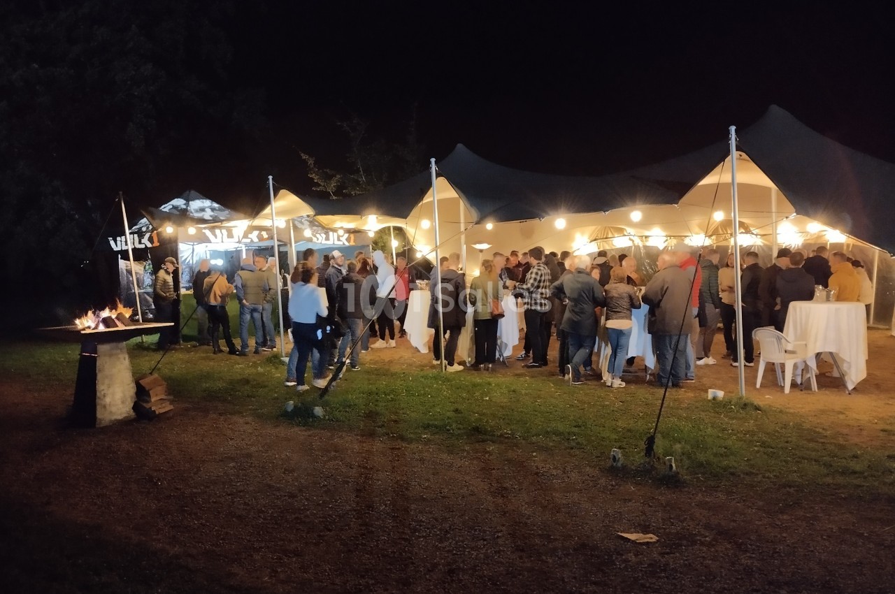 Groupe de personnes rassemblées sous des tentes éclairées en soirée, avec un brasero allumé à gauche.
