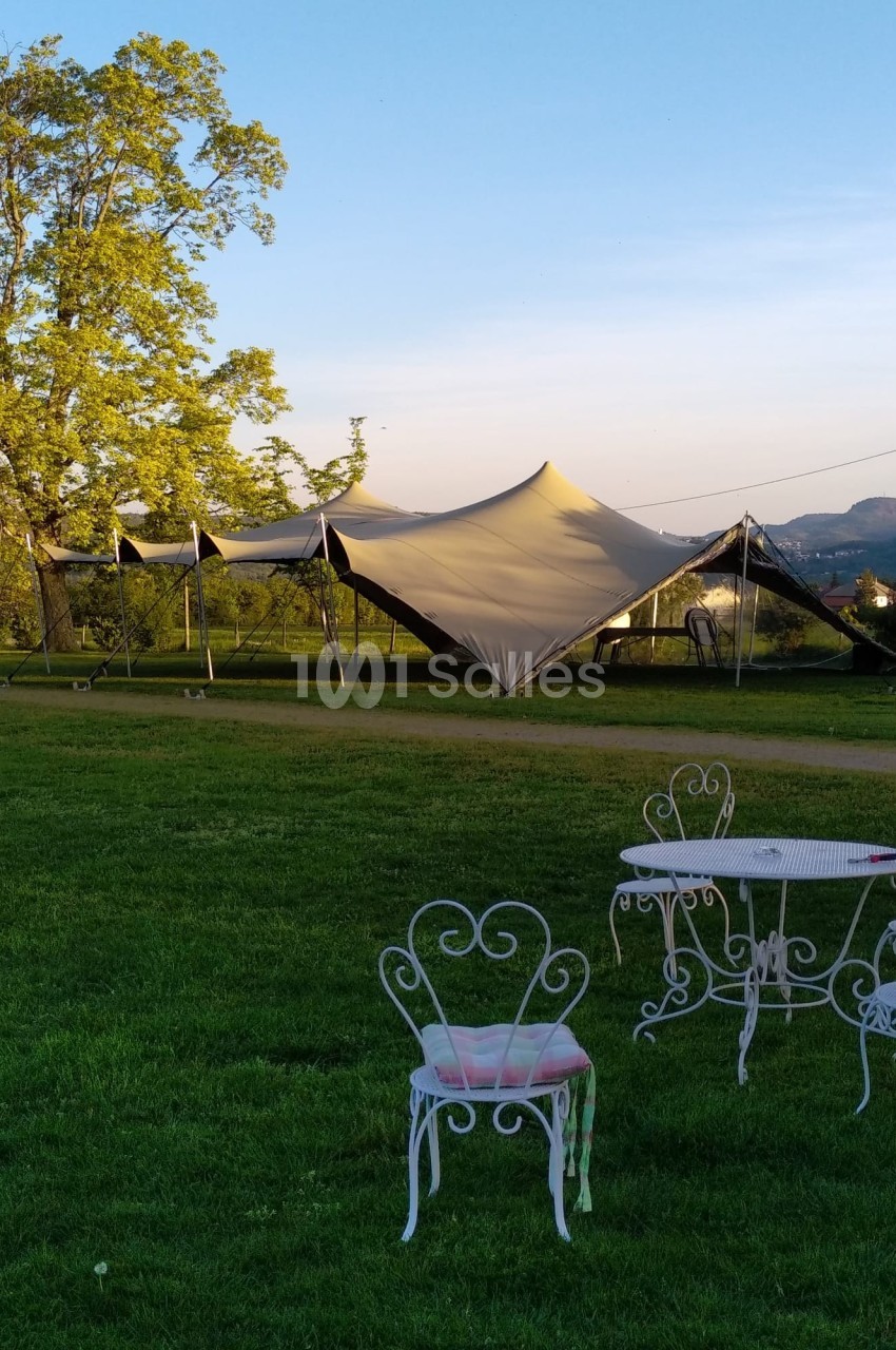 Tente beige installée sur une pelouse verdoyante avec des chaises et une table en fer forgé au premier plan.
