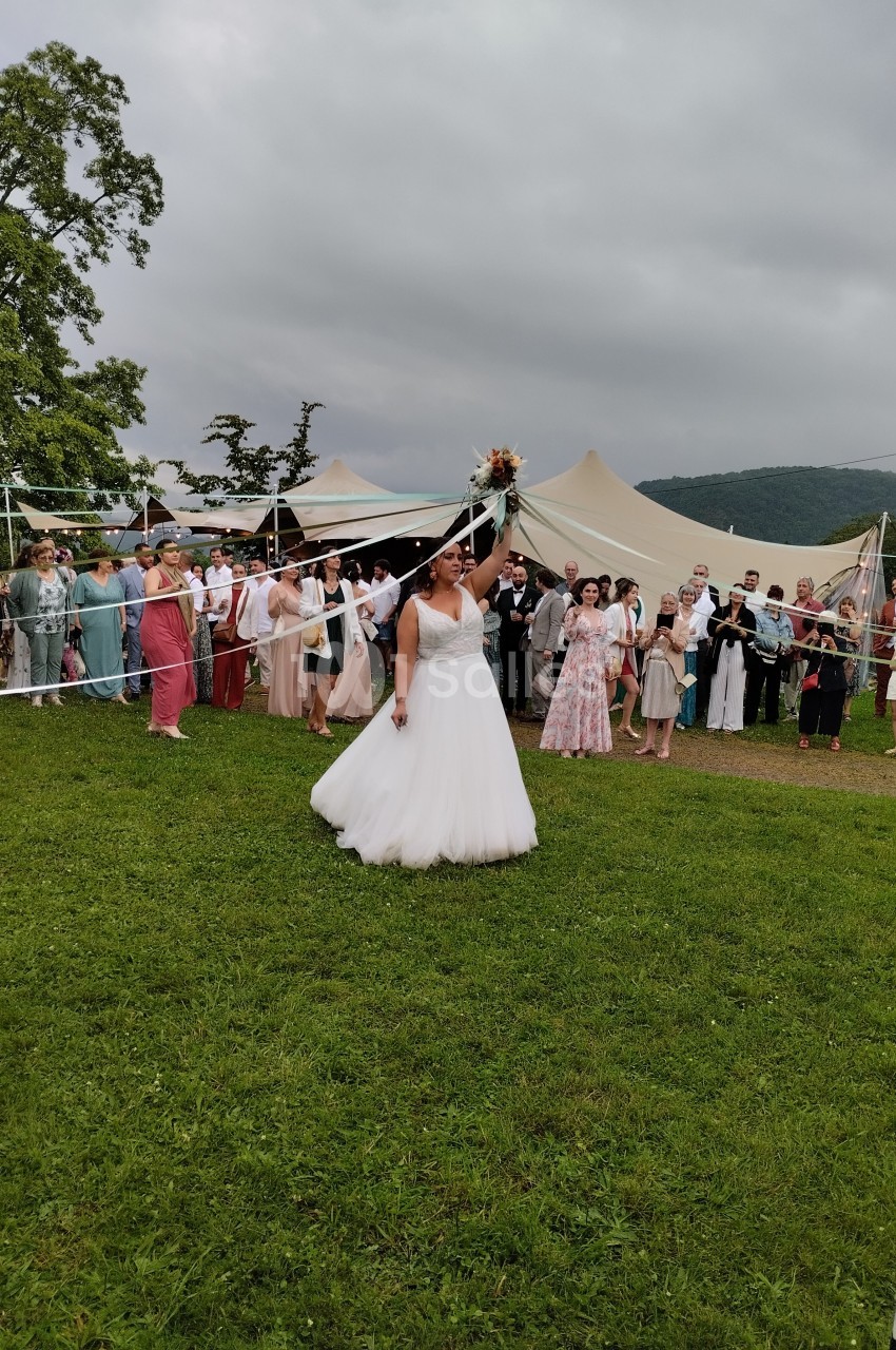 Une mariée en robe blanche lance son bouquet devant un groupe de convives dans un cadre extérieur verdoyant.