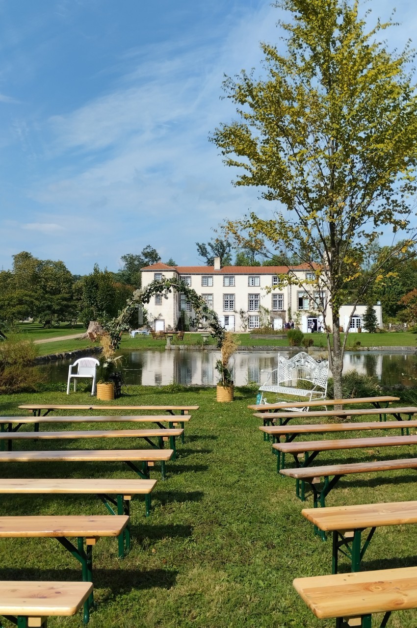 Bancs en bois disposés en rangées sur une pelouse face à un étang et une grande maison blanche entourée de verdure.