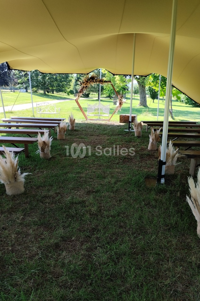 Allée centrale d'une cérémonie en plein air sous une tente, décorée de bancs en bois et de bouquets de pampas.