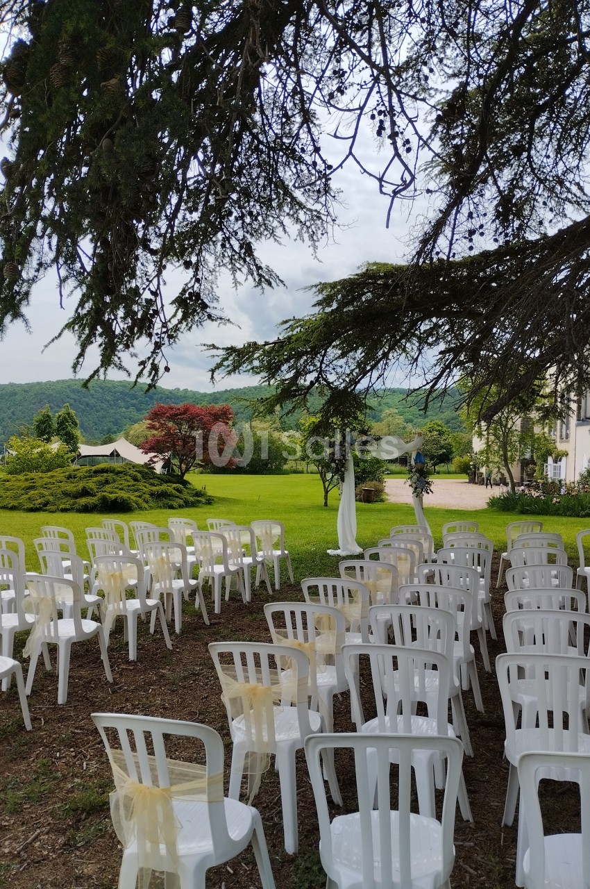 Chaises blanches disposées en rangées sous un arbre, face à un espace vert avec une tente et des collines en arrière-plan.