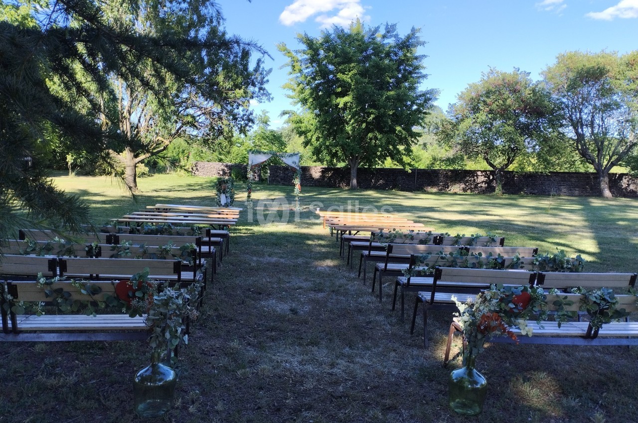 Allée centrale bordée de bancs décorés de feuillages, menant à une arche de mariage dans un jardin verdoyant.