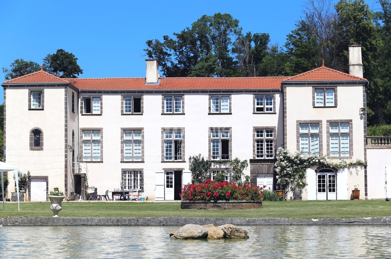 Grande maison blanche à trois étages avec toit en tuiles rouges, entourée de verdure et située près d'un plan d'eau.