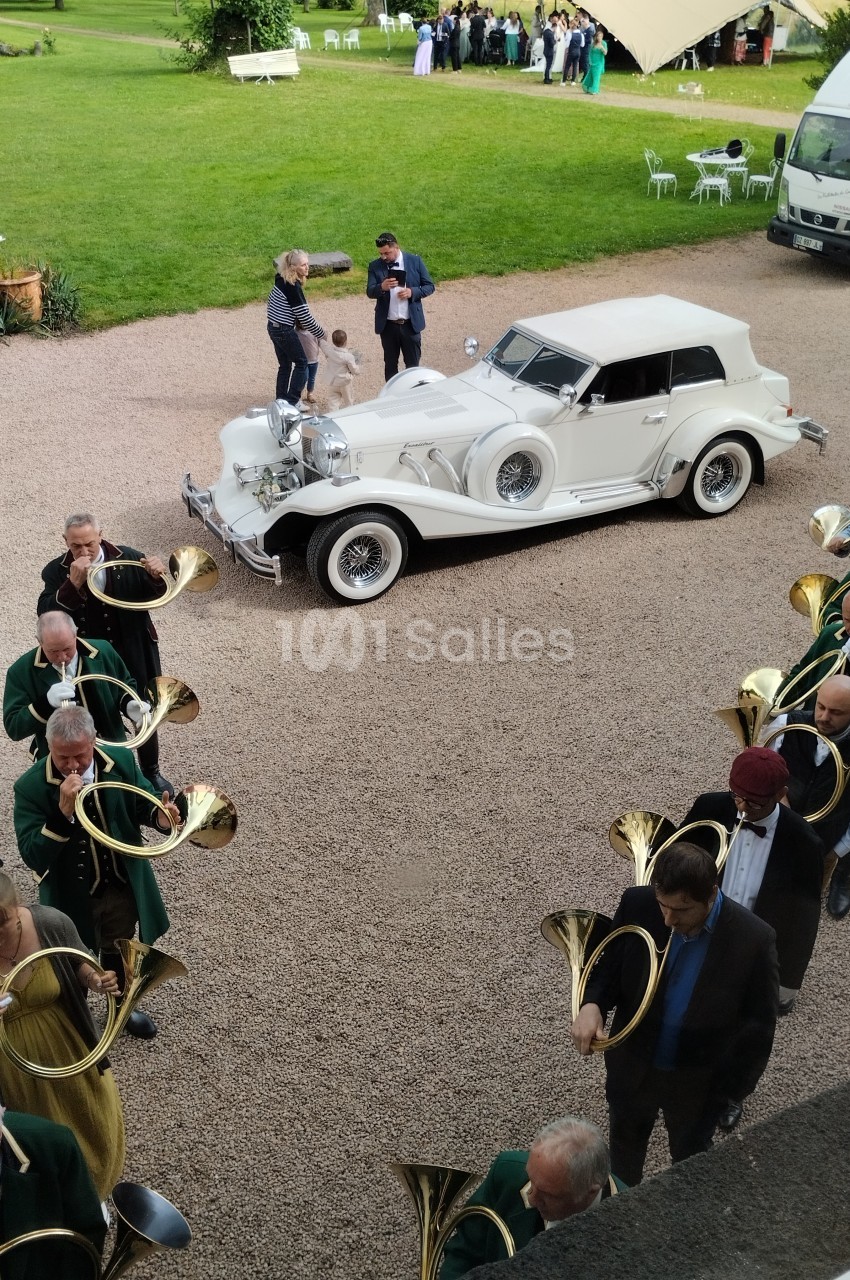 Un groupe de musiciens jouant du cor autour d'une voiture ancienne blanche garée sur une allée gravillonnée.