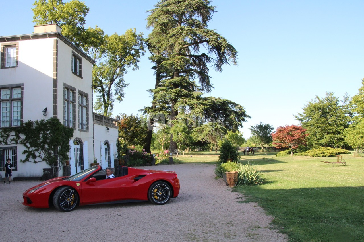 Voiture rouge décapotable garée devant une maison blanche entourée de verdure et d'arbres sous un ciel dégagé.