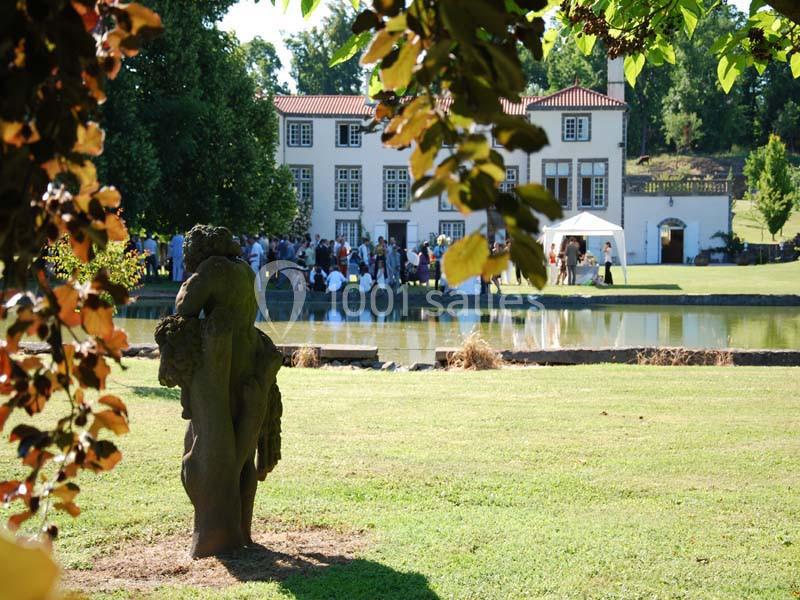 Statue en pierre dans un parc verdoyant, avec un bâtiment historique et des personnes rassemblées en arrière-plan.