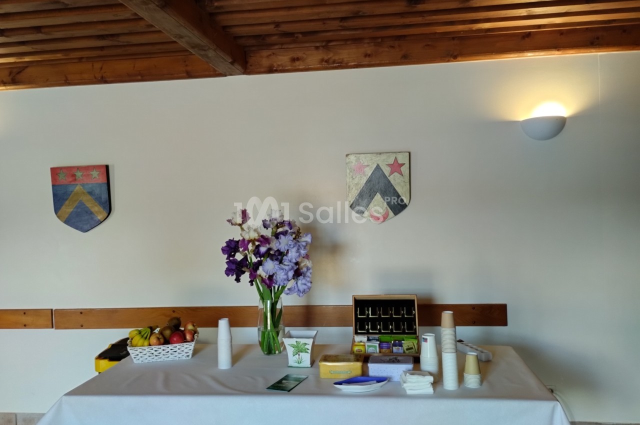 Table dressée avec des fleurs, fruits, boissons et accessoires, dans une salle avec des blasons accrochés au mur.