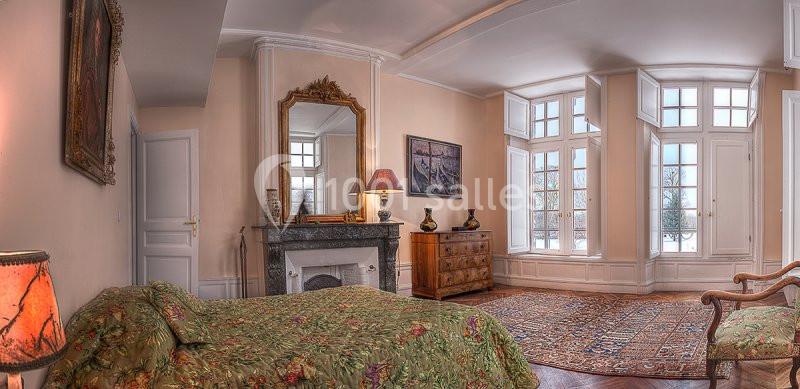 Chambre lumineuse avec lit à motif floral, cheminée ornée d'un miroir, commode en bois et grandes fenêtres donnant sur l…