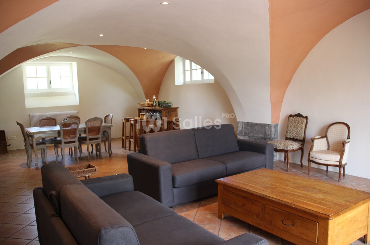 Salle voûtée avec canapés gris, table basse en bois, table à manger et chaises anciennes sur un sol en carrelage.