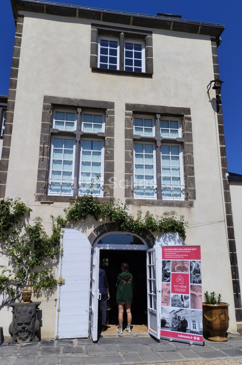 Façade d'un bâtiment en pierre avec une porte ouverte, des visiteurs entrant et un panneau d'exposition à droite.