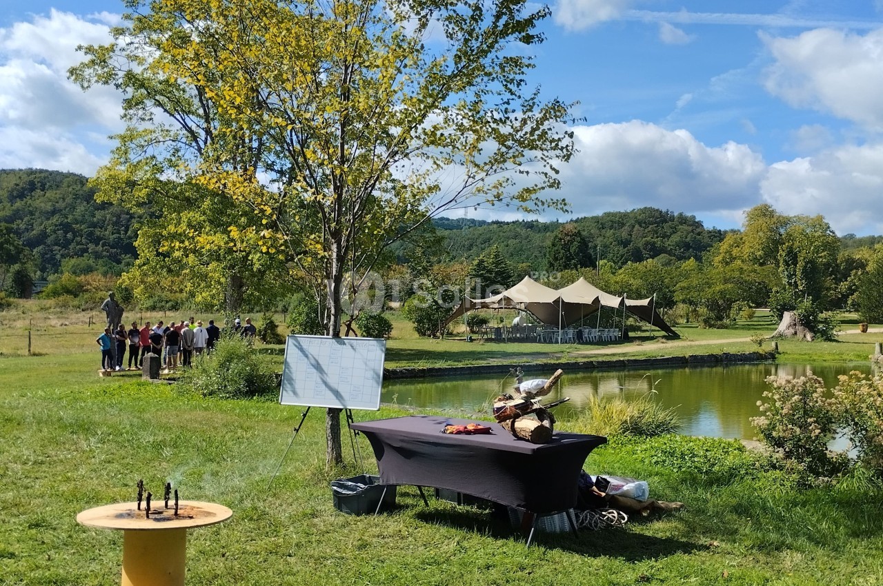 Un espace extérieur avec une table dressée près d'un étang, des arbres et un groupe de personnes au loin.