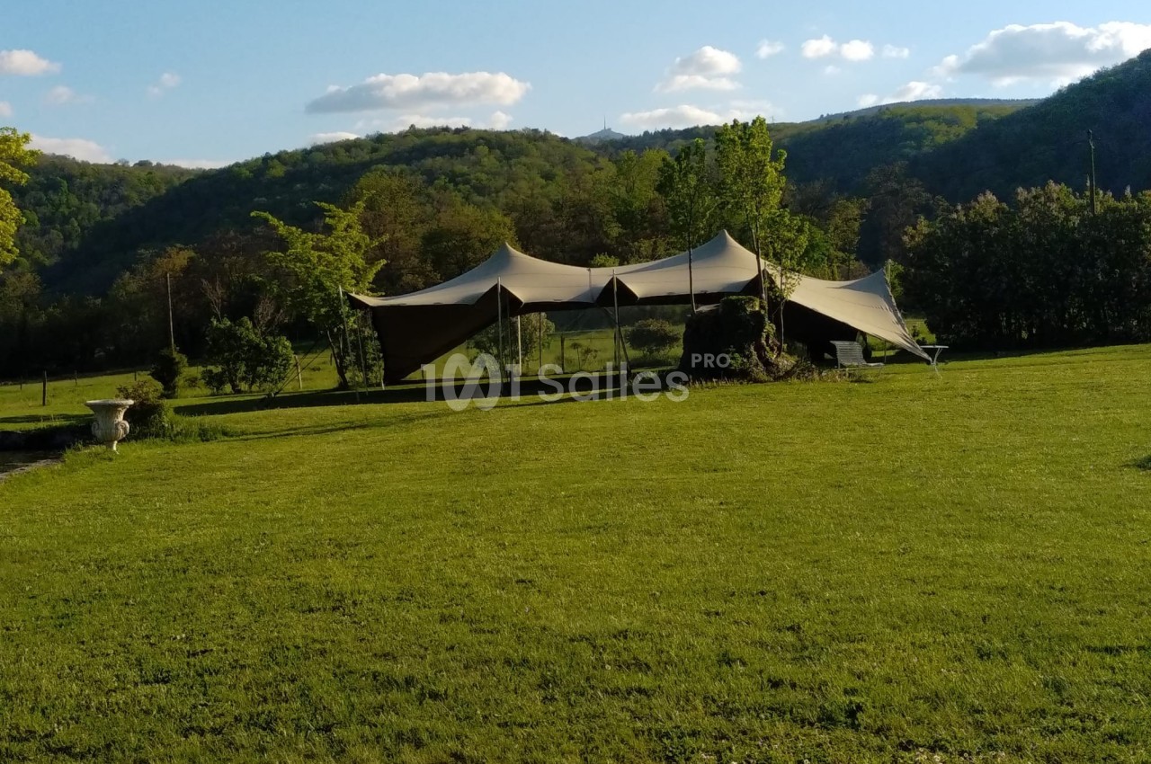 Tente beige installée sur une pelouse verdoyante, entourée de collines et d'arbres sous un ciel partiellement nuageux.
