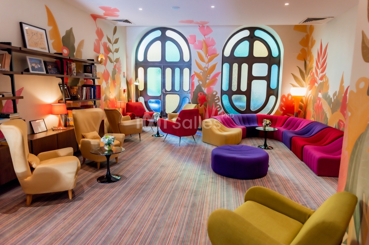 Salon coloré avec fauteuils et canapés modernes, grandes fenêtres arrondies et décor mural floral.