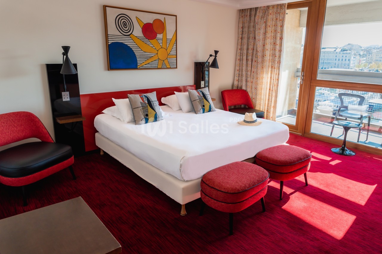 Chambre d'hôtel lumineuse avec lit double, fauteuils rouges, tableau coloré et balcon donnant sur une vue urbaine.