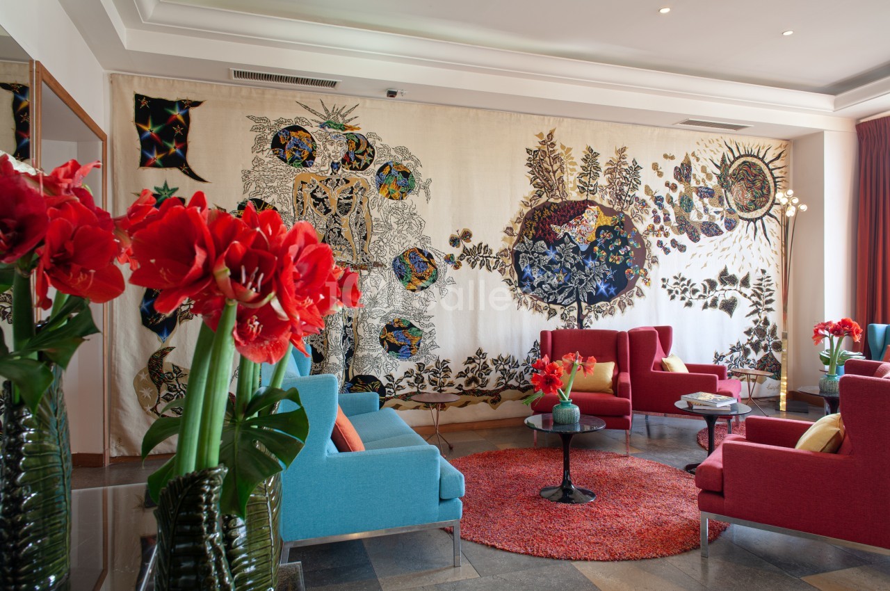 Salon lumineux avec fauteuils colorés, tapis rouge, fleurs rouges en vase et une grande tapisserie murale décorative.