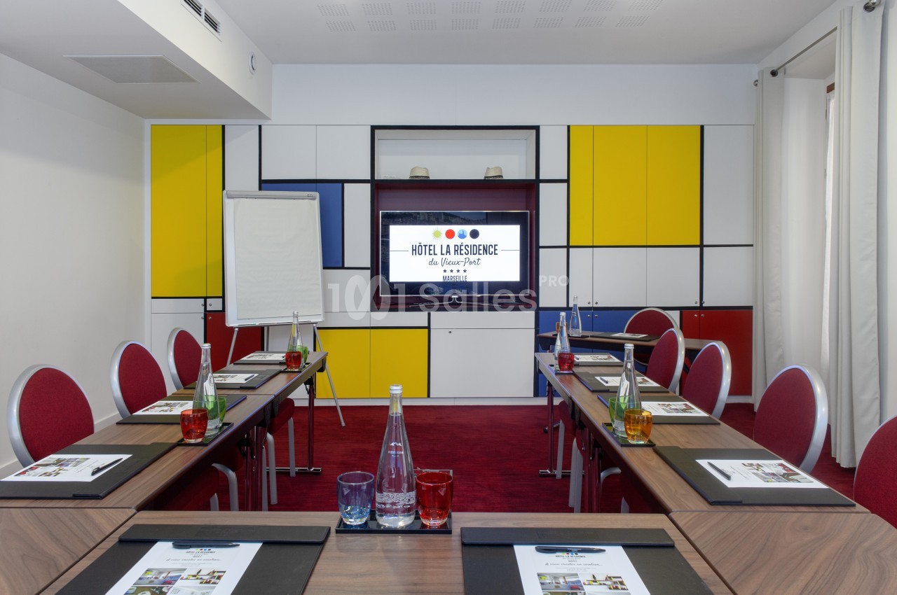 Salle de réunion avec tables disposées en U, chaises rouges, écran affichant un logo, et décor coloré en arrière-plan.