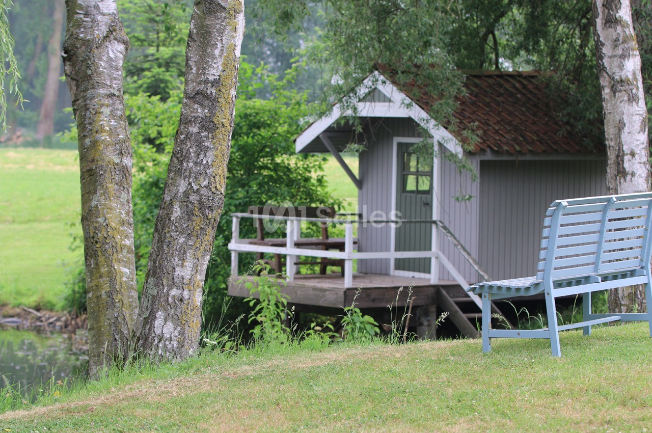 Petite cabane en bois avec terrasse au bord d'un étang, entourée d'arbres et d'une pelouse avec un banc.