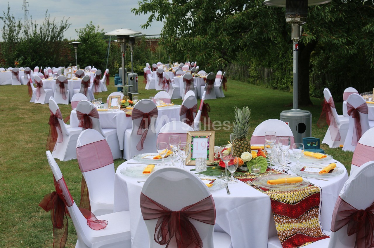 Tables rondes décorées avec nappes blanches et rubans rouges, disposées en extérieur pour un événement festif.