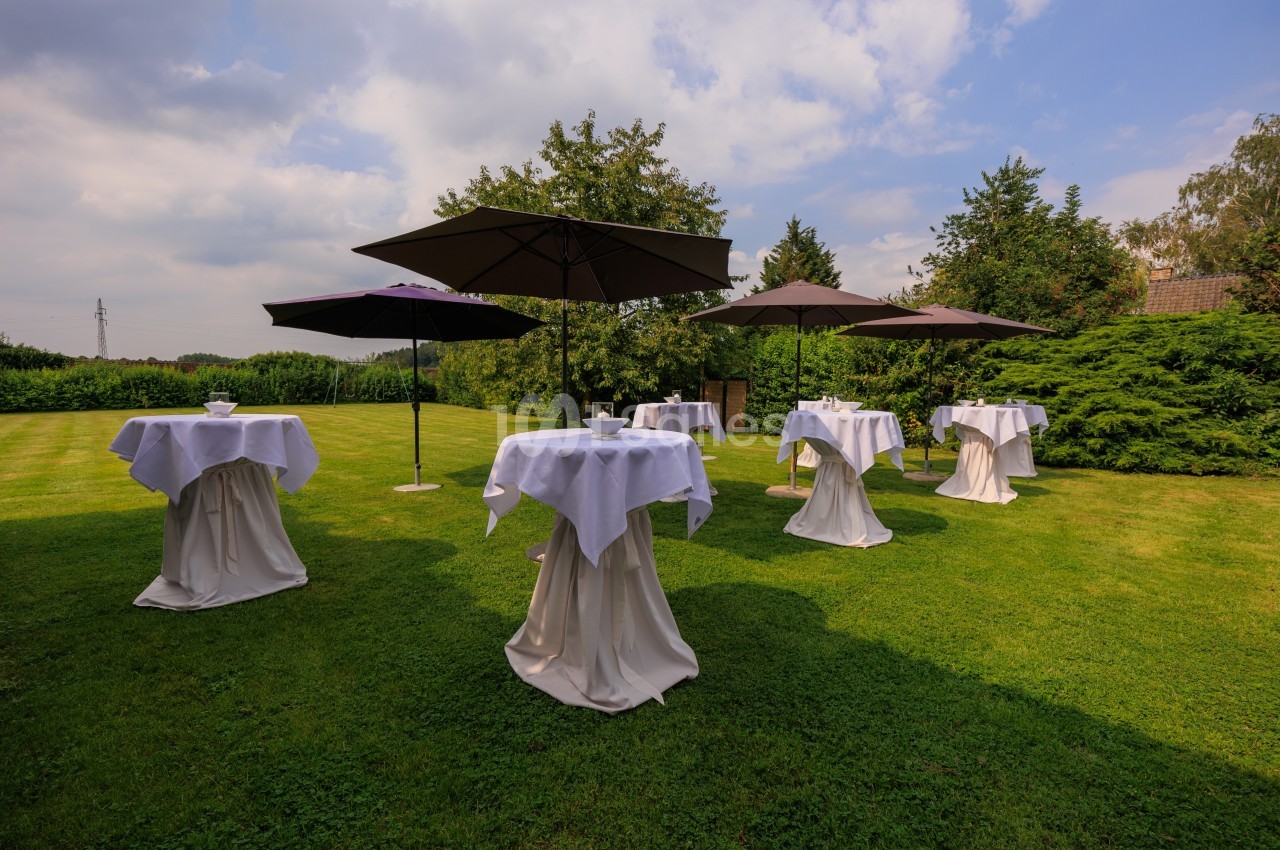 Tables hautes drapées de nappes blanches avec parasols noirs sur une pelouse, entourées de verdure et d'arbres.