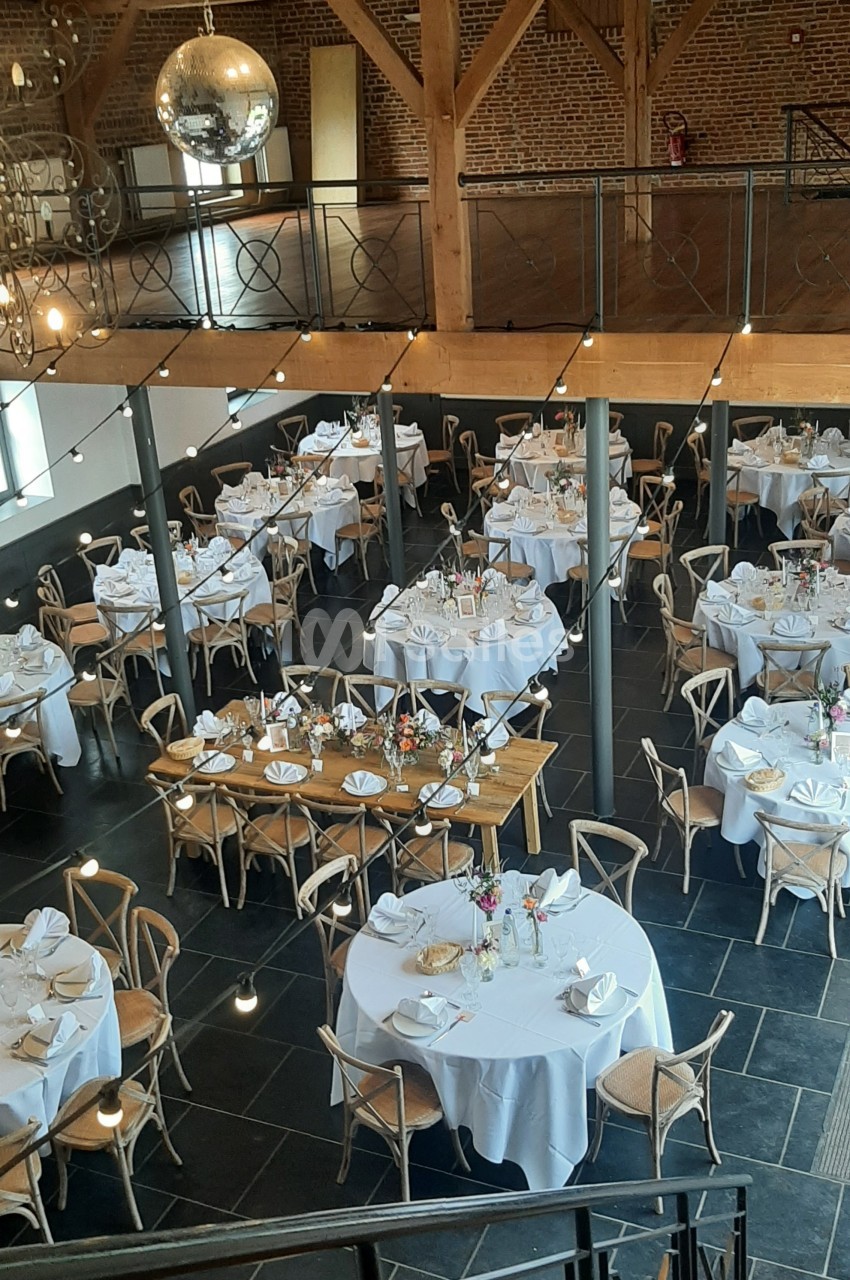 Salle de réception élégante avec tables rondes et rectangulaires dressées, décorée de fleurs et éclairée par une boule…