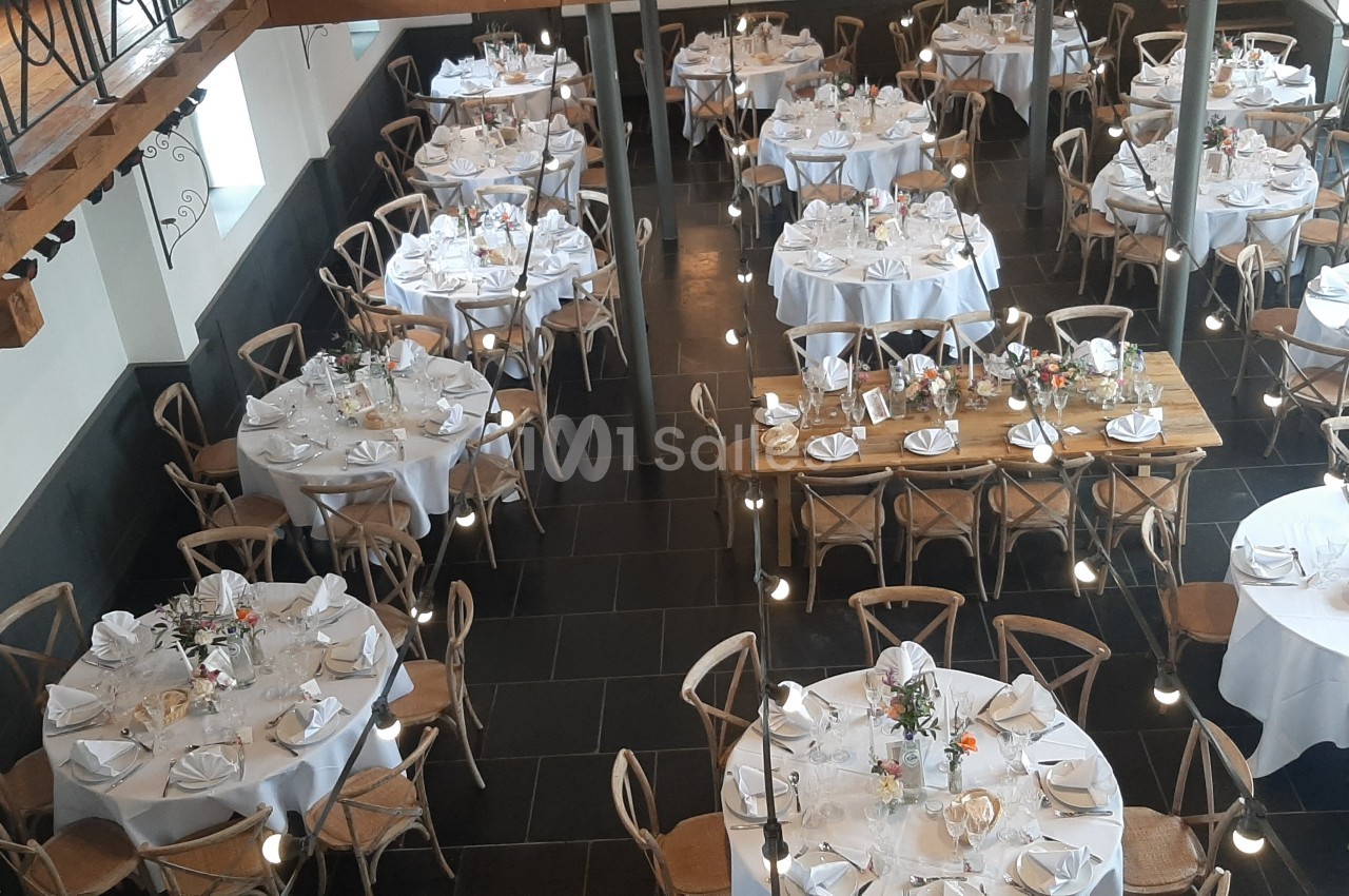 Salle de réception avec tables rondes et rectangulaires dressées, nappes blanches et décorations florales.