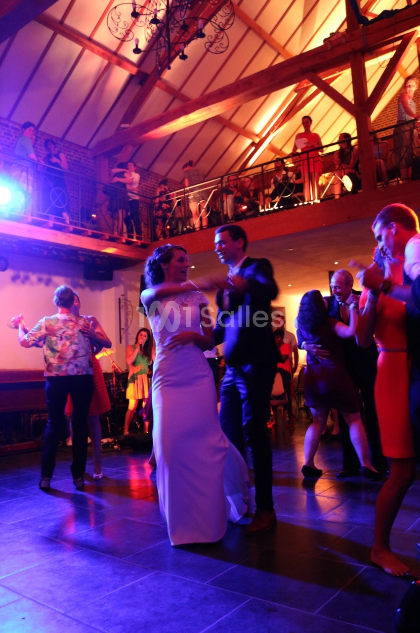 Un couple danse au centre d'une salle de réception éclairée, entouré d'invités sur la piste et à l'étage.