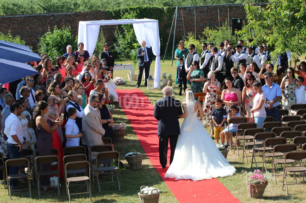Un mariage en plein air avec une mariée marchant sur un tapis rouge, accompagnée d'un homme, devant des invités assis.