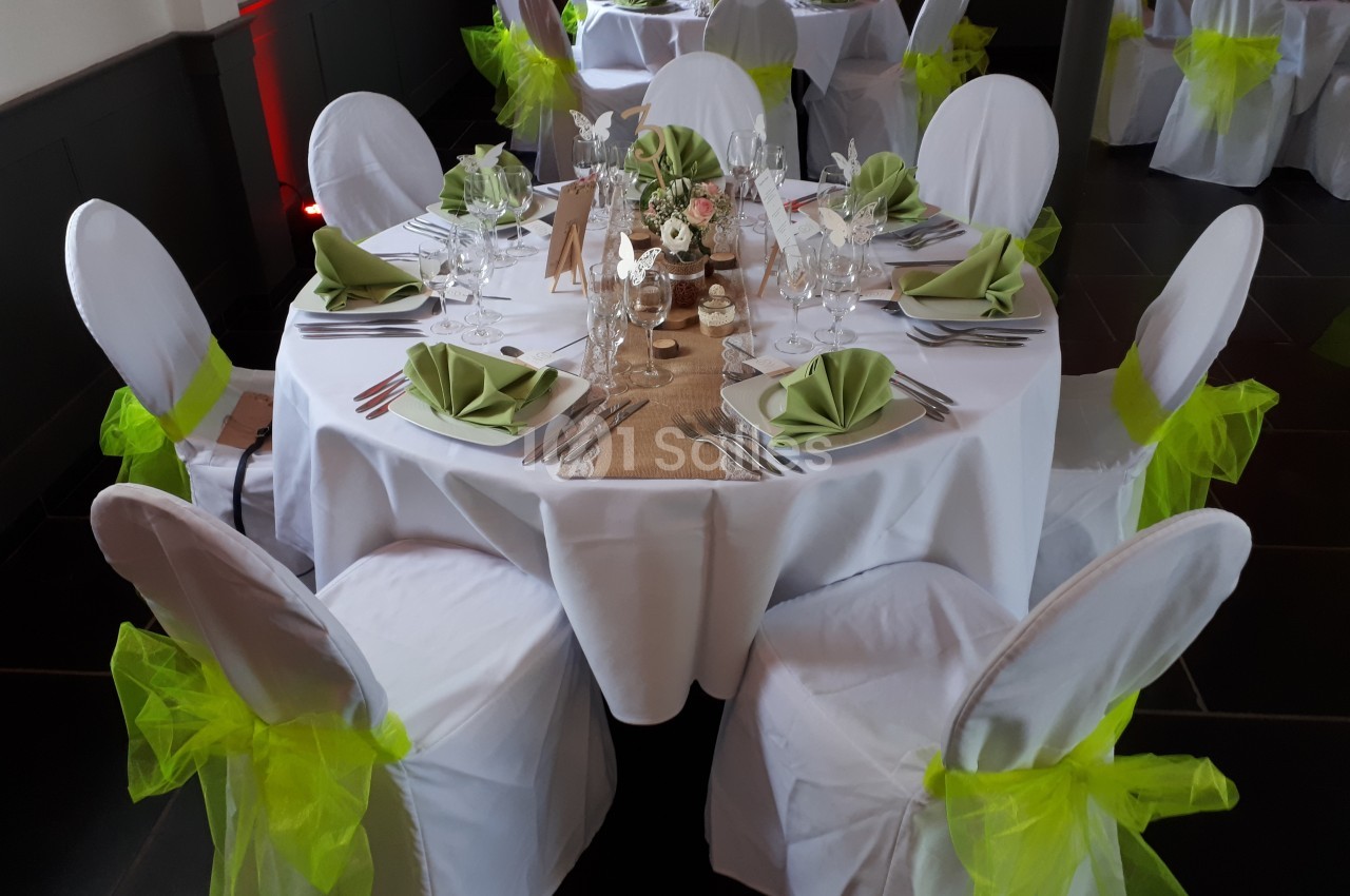 Table de réception décorée avec nappes blanches, serviettes vertes pliées, chaises ornées de nœuds verts et centre de table…