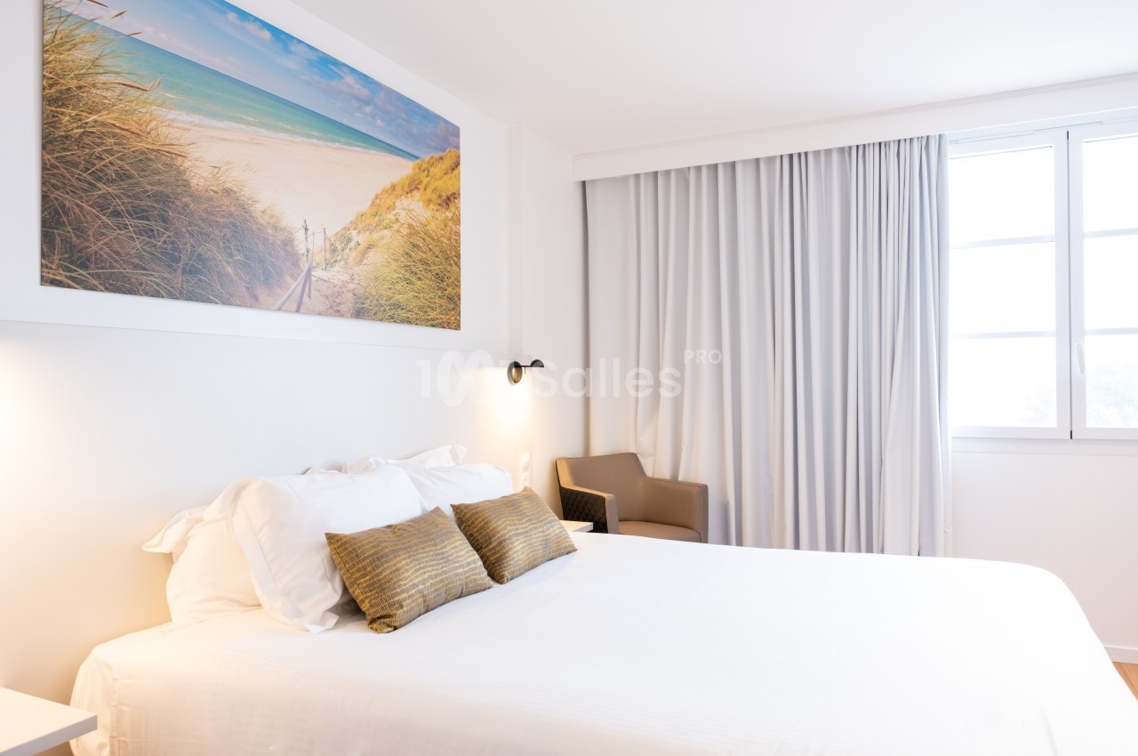 Chambre lumineuse avec un lit double, des coussins dorés, un fauteuil beige et un tableau représentant une plage.
