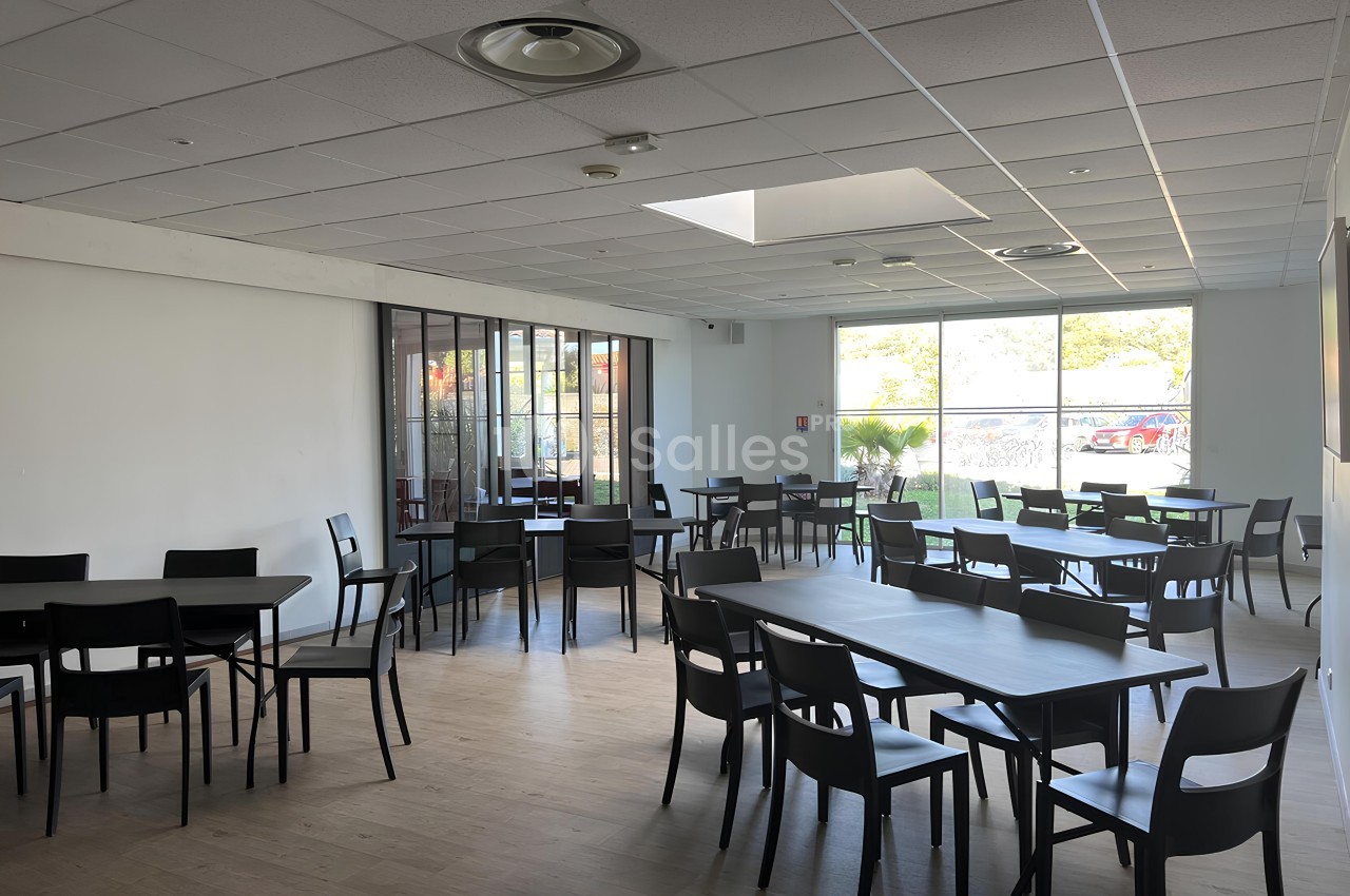 Salle lumineuse avec tables et chaises noires, grandes fenêtres laissant entrer la lumière naturelle.