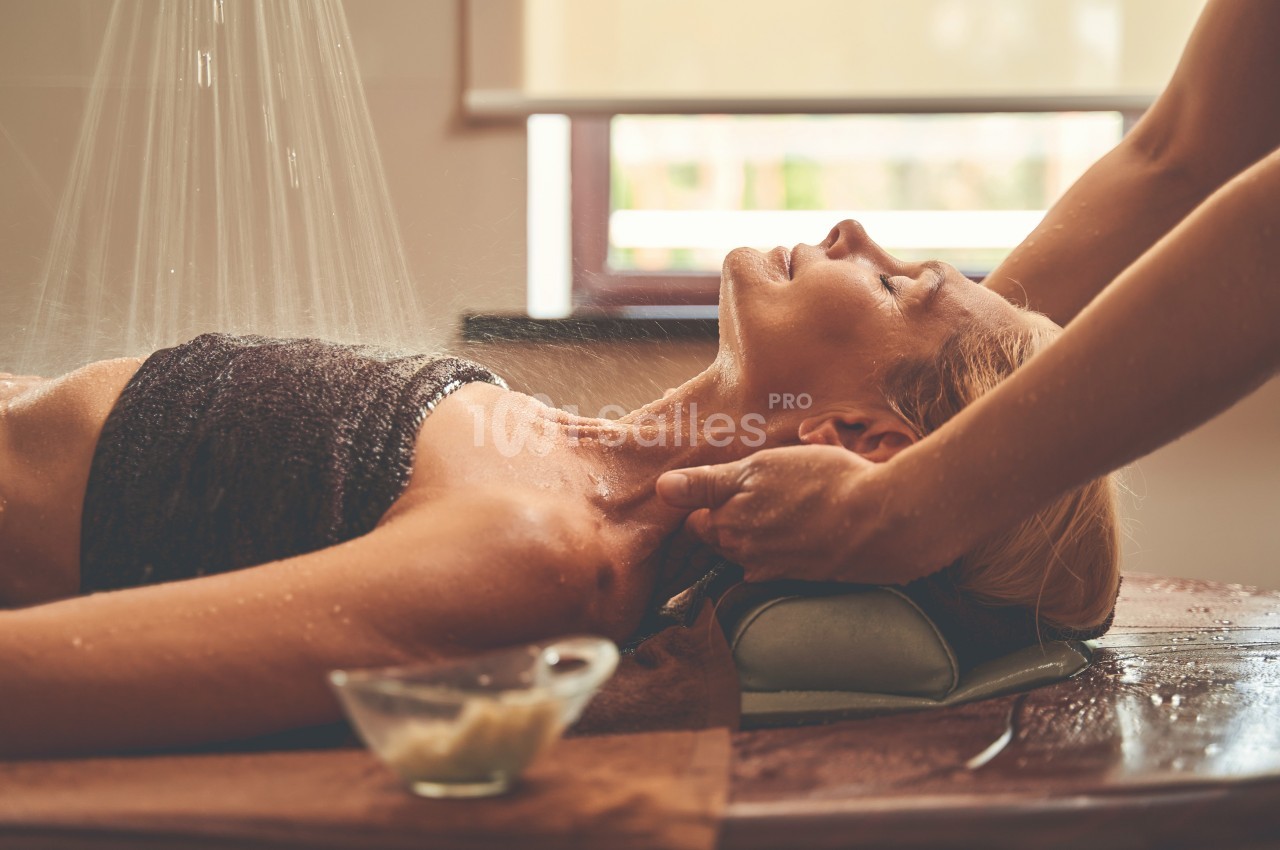 Femme allongée recevant un massage sous une douche d'eau, dans un cadre lumineux et relaxant.