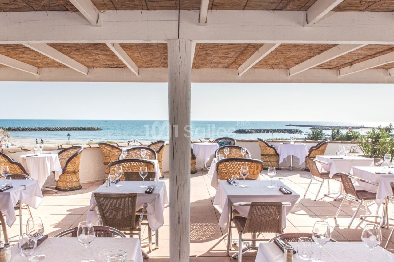 Terrasse d'un restaurant en bord de mer avec des tables dressées, vue sur la plage et l'océan en arrière-plan.