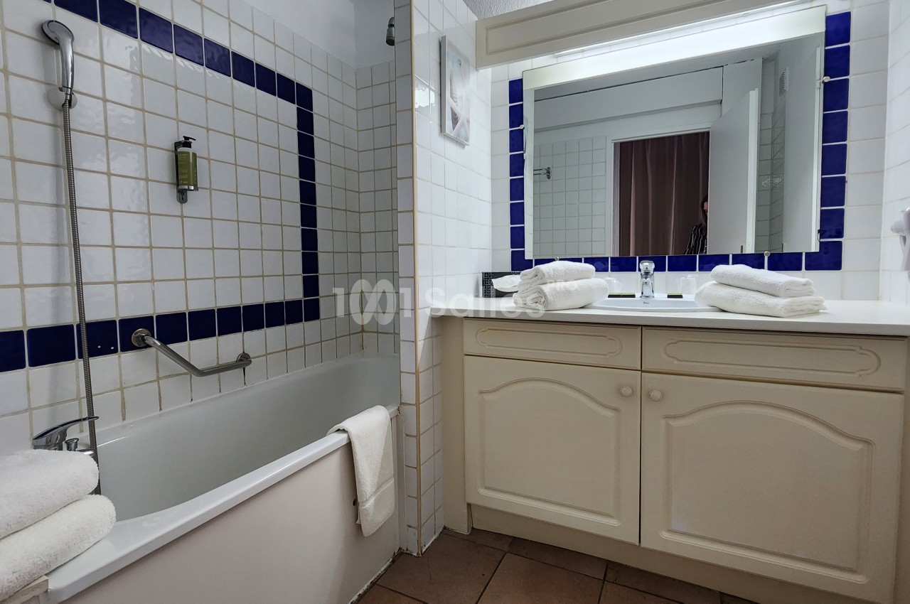 Salle de bain avec baignoire, carrelage blanc et bleu, lavabo avec miroir éclairé, serviettes pliées sur le plan de travail.